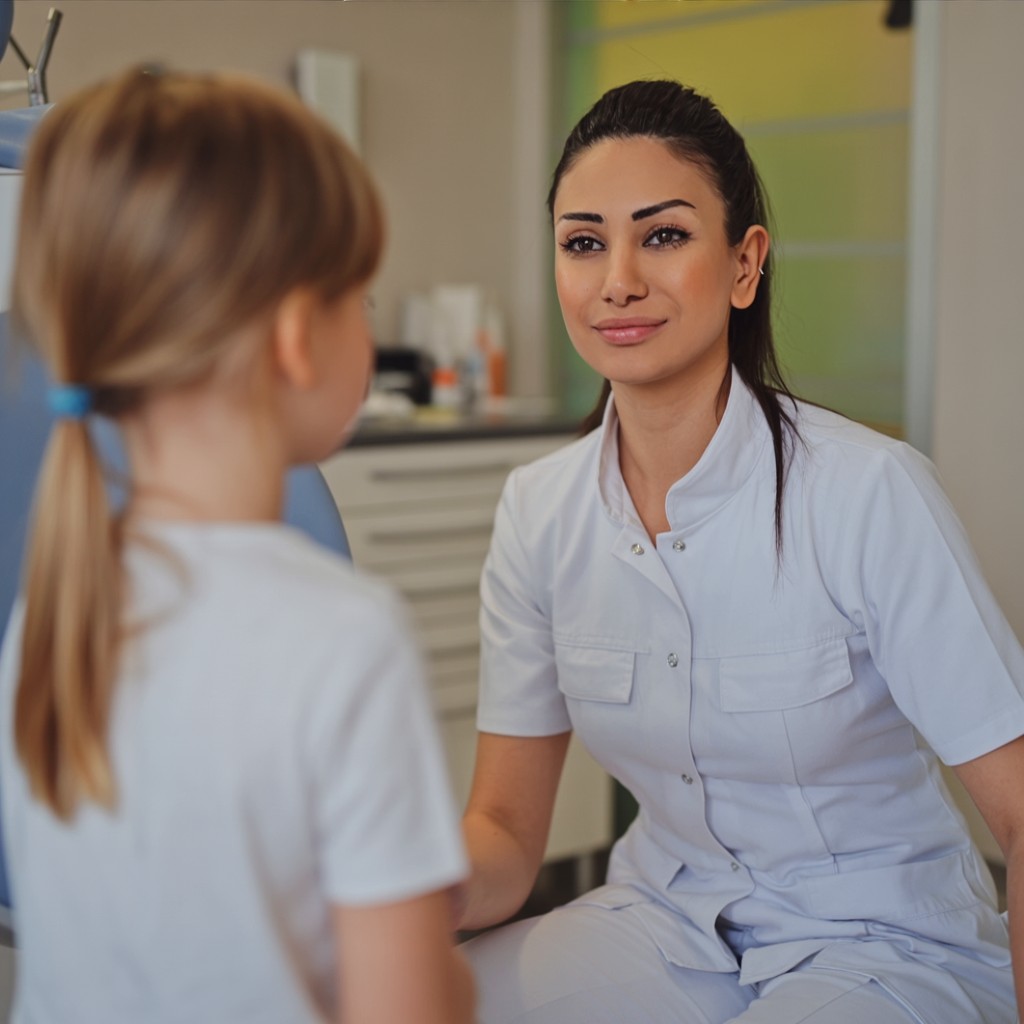"Tandarts in wit uniform praat met een meisje in een tandartspraktijk, met een knuffeldinosaurus op de tafel."